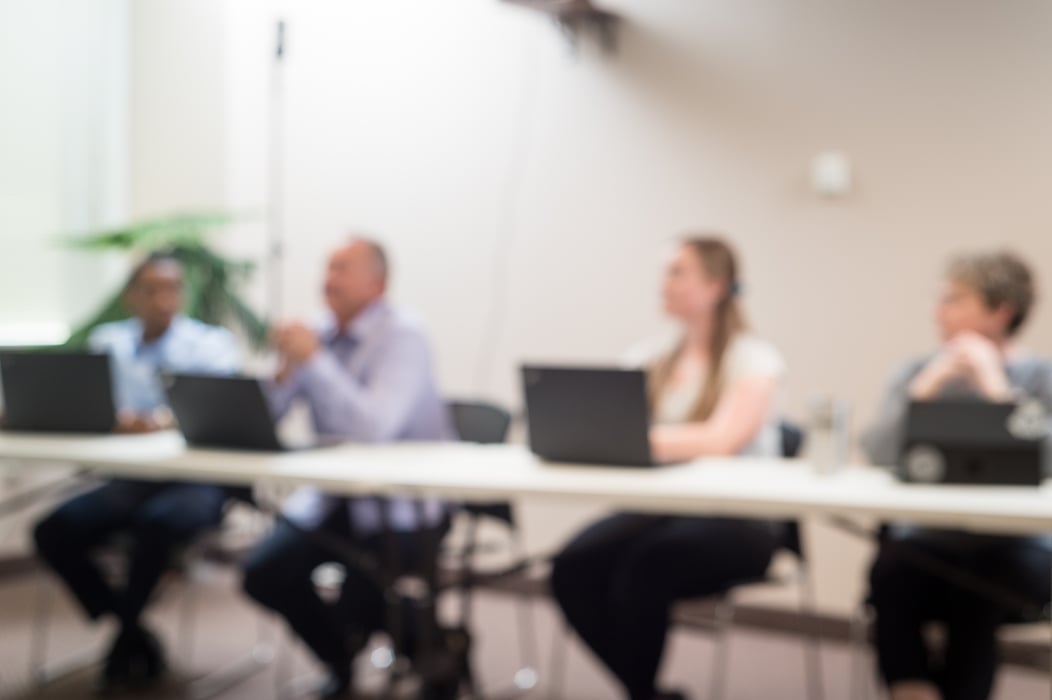 A blurred image of 4 people sitting next to each other at a table with laptops in front of them looking off screen.